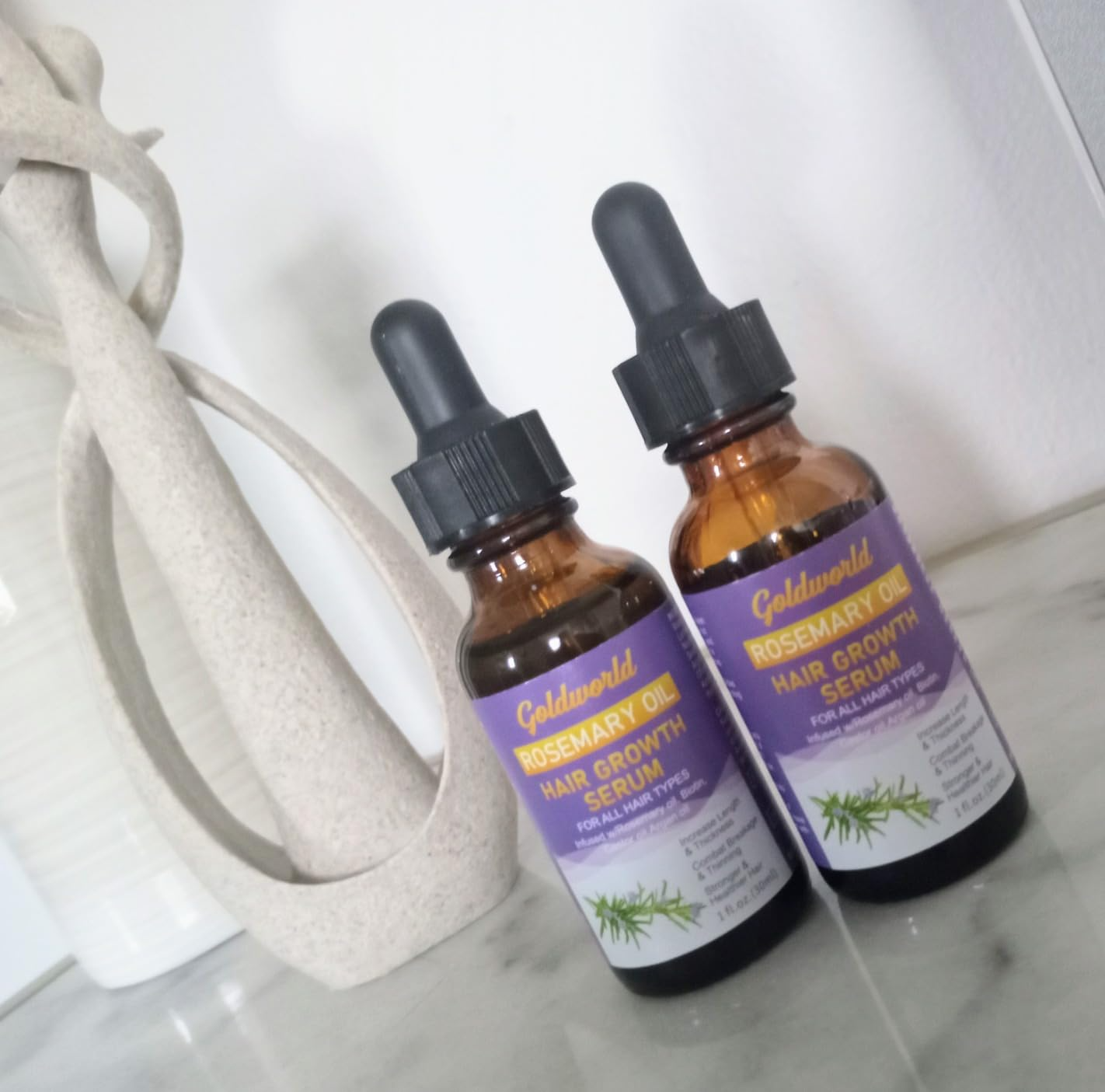Two bottles of Goldworld Rosemary Oil Hair Growth Serum on marble surface. Natural hair care product display.