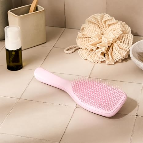 Pink detangling hairbrush on a bathroom counter with oil bottle and bath sponge in the background