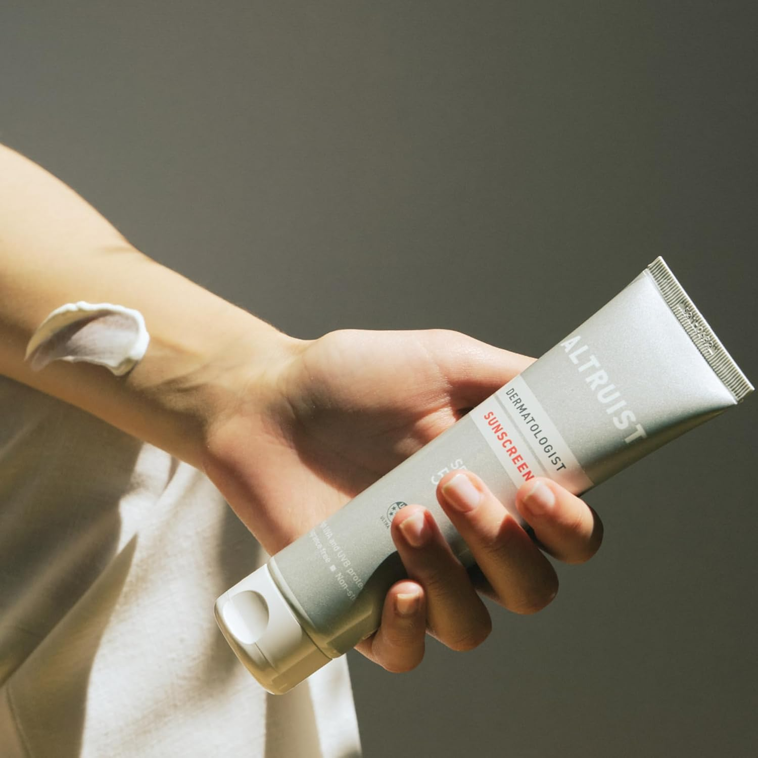 Hand holding Altruist Dermatologist Sunscreen tube with lotion applied on forearm, highlighting skincare and sun protection