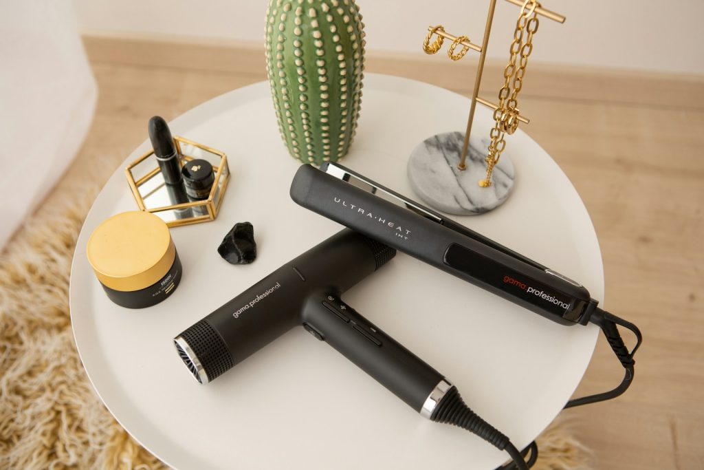 Hair styling tools and beauty products on a white table, featuring a hairdryer and flat iron near jewelry and cosmetics