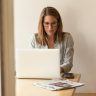 Woman working on laptop with glasses, sitting at a desk with newspapers and phone, focused on tasks