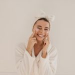 woman in a bathrobe smiles while gently holding her face, conveying a sense of joy and relaxation