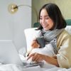 Woman working on a laptop in bed, smiling and holding a mug, cosy morning atmosphere