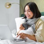 Woman working on a laptop in bed, smiling and holding a mug, cosy morning atmosphere