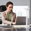 A woman in a business suit sits at a desk, working on a laptop in a professional office setting