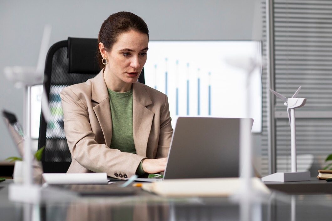 A woman in a business suit sits at a desk, working on a laptop in a professional office setting