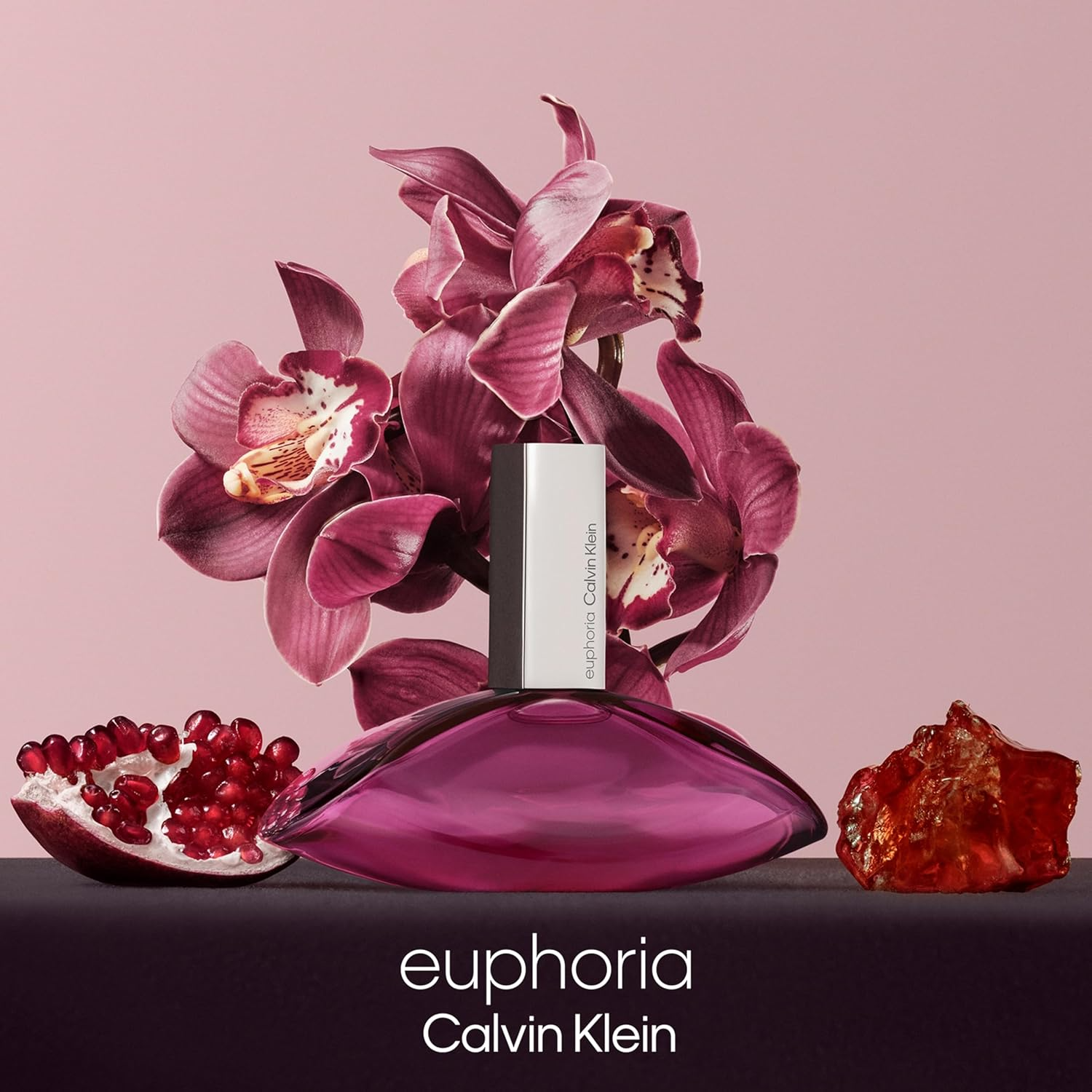 Calvin Klein Euphoria perfume with pink orchids, pomegranate, and amber gemstone on a soft pink background